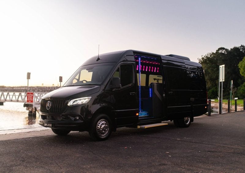 Black 20 seat party limo in Sydney, a Mercedes Sprinter parked by the water at dusk with the side door open showing pink and blue LED interior lighting